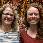 Erin and Joanna smile in front of a blueberry bush.