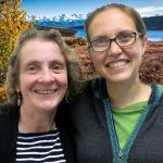 Christa and katie smile at the camera in front of a lake with mountains in the distance.