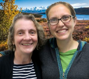 Christa and katie smile at the camera in front of a lake with mountains in the distance.