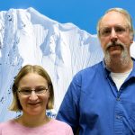 An older man and younger woman look at the camera. Behind them a wall of snow rises.