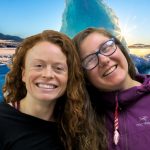 Tow women lean their heads together in front of a blue iceberg.