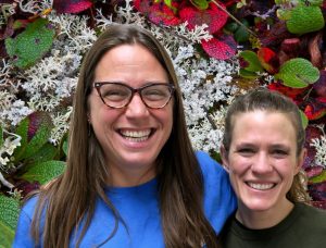 Jessica and Jessie smile in front of a bed of lichen and leaves.