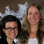 Two women smile in front of a close-up picture of a snowflakeʼs ice crystals.