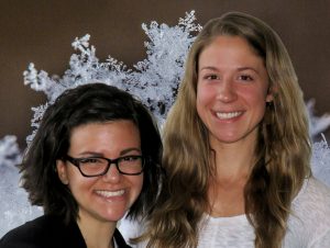 Two women smile in front of a close-up picture of a snowflakeʼs ice crystals.