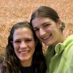 Jessiea nd Kristin smile at the camera, a field of grain behind them.