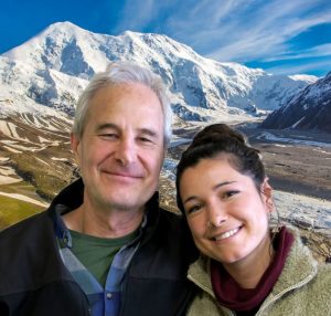 Rick and Naomi put their heads together and smile in front of a mountain and streams.