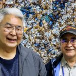 Orville and Justin, two older men with glasses, smile in front of kelp and clam beds.