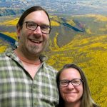 Ryan and Holly with their arms around each other smile in front of an aerial view of a forest.