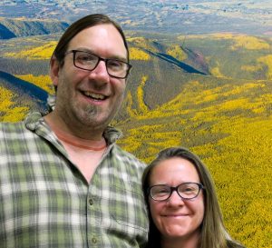 Ryan and Holly with their arms around each other smile in front of an aerial view of a forest.