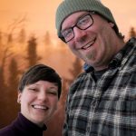 Erica, a young woman with short brown hair, and Ryan, a man with a goatee and glasses, smile in front of an orange sunset sky.