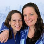 Audrey and Shannon, two women who look similar with dark brown hair have their arms around eachother in front of snowy trees.