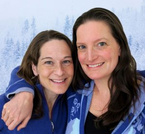 Audrey and Shannon, two women who look similar with dark brown hair have their arms around eachother in front of snowy trees.