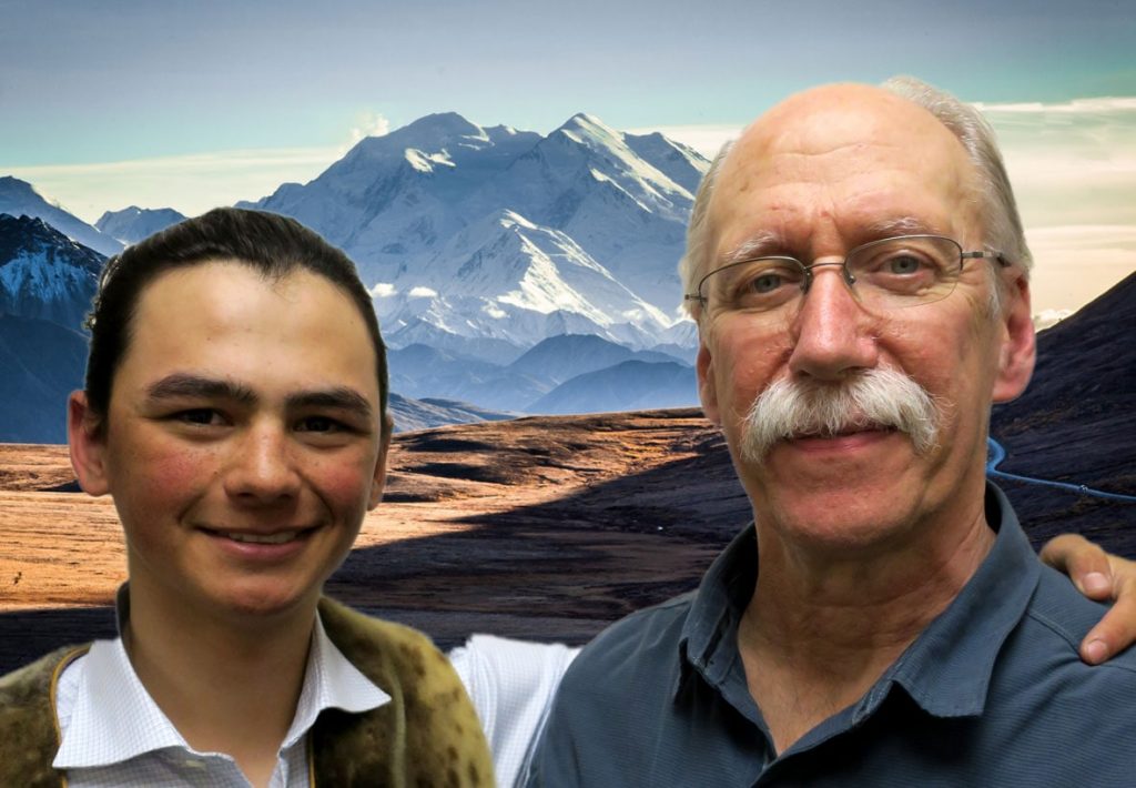 Sam, a boy with dark hair and freckles, and Luke an older man with a white mustache, smile in front of Denali.