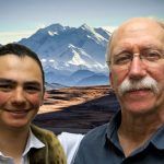 Sam, a boy with dark hair and freckles, and Luke an older man with a white mustache, smile in front of Denali.