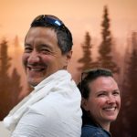 Arlo and Jessica stand back to back and smile in front of trees and an orange sky.