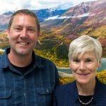 Kathy and Matt smile at the camera in front of a photoshopped background of a river shrub-covered mountains.