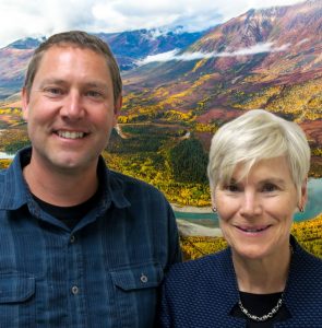 Kathy and Matt smile at the camera in front of a photoshopped background of a river shrub-covered mountains.