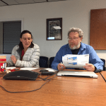 Rick Thoman and Gay Sheffield face the camera sitting at a table. They're sitting in front of a conference call phone and rick is holding a briefing report paper.