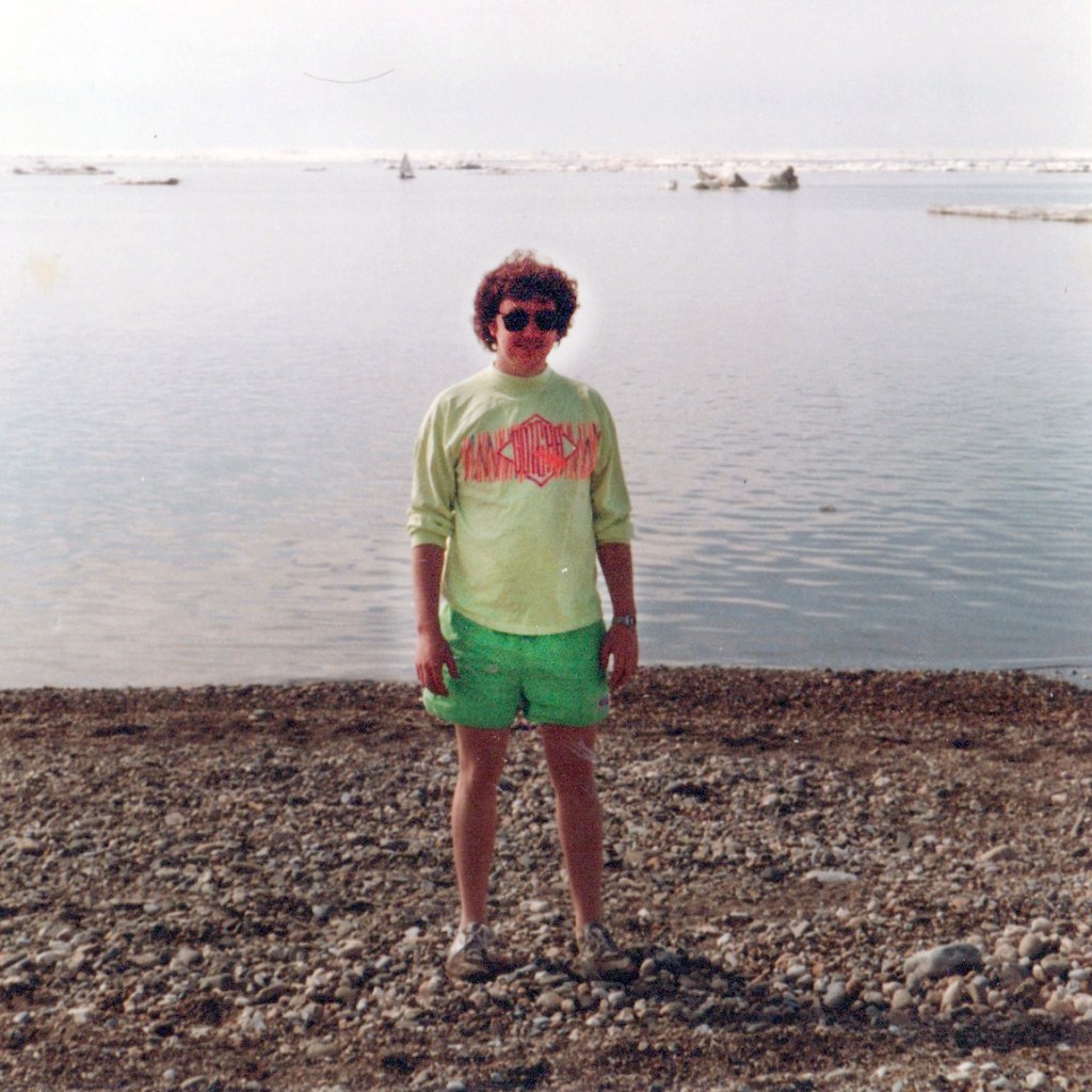 A young Rick Thoman smiles at the camera in an older, grainy photo on of a rocky beach. Small ice bergs float in the water behind him.