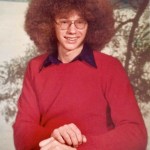 A young Rick Thoman in a red sweater and large afro hair style smiles into the camera in a high school photo style potrait.