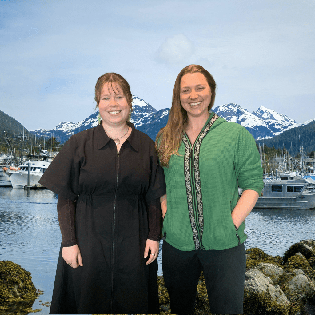 Two women smile in front of a photoshopped background of a Sitka marina. 