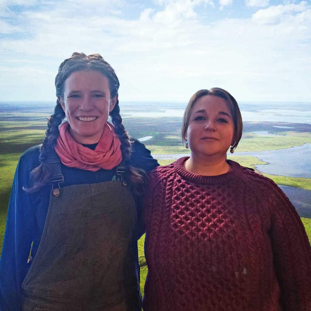 Two women smile in front of a photoshopped background of a vast swampy tundra. 