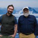 Two men, one with a white beard cap and the other with a brown beard and hair stand and smile in front of a photoshopped background of icy mountains.