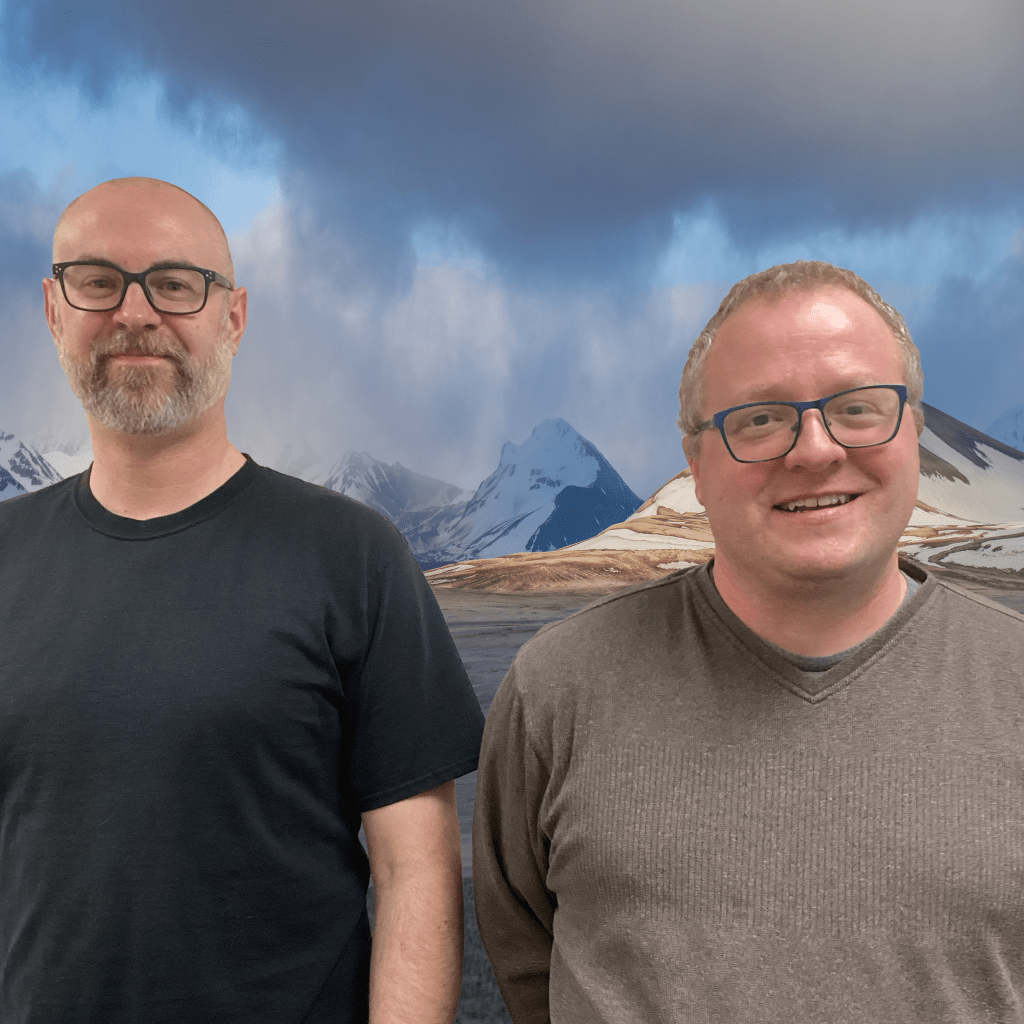 Two men wearing similar glasses and t-shirts smile in front of a photoshopped background of the Mount Katmai volcano.