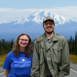 Two young adults smile in front of a photoshopped background of a road leading to Denali.