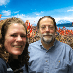 A woman and man in smile in a selfie in front of a photoshopped background of blueberry bushes and mountains int he distance.