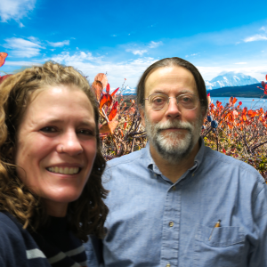 A woman and man in smile in a selfie in front of a photoshopped background of blueberry bushes and mountains int he distance.