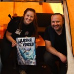 A woman and man smile while crouching inside a yellow Arctic Oven tent. The woman holds an Alaska Voices poster.
