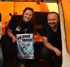 A woman and man smile while crouching inside a yellow Arctic Oven tent. The woman holds an Alaska Voices poster.