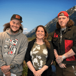 Three Native Southeast Alaskan people, two men and a woman, smile in front of a photoshopped background of a rocky moutain.