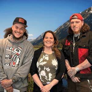 Three Native Southeast Alaskan people, two men and a woman, smile in front of a photoshopped background of a rocky moutain.