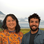 A Native Alaskan woman and and a man with dark hair smile at the camera in front of a photshopped background of mountains.