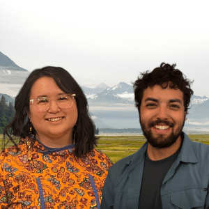 A Native Alaskan woman and and a man with dark hair smile at the camera in front of a photshopped background of mountains.